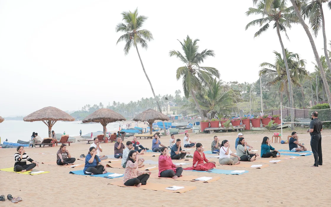 A class seated on mats on the beach under the palms at The Leela Kovalam, Abhilash standing to one side.